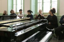 Seitliche Ansicht eines Klaviers mit Musikschülerinnen und -schülern im Hintergrund.